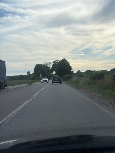 The image depicts a road scene, likely in Kumachevo, Russia, with a cloudy sky overhead. In the foreground, the hood of a vehicle is visible, suggesting the photo was taken from inside a car. The road stretches into the distance, with a white line marking the lane. On the left side of the road, a large blue truck is partially visible, with its wheels and side in view. Further down the road, two cars are traveling in the same direction. The first is a white sedan, and behind it is a dark-colored SUV. The surrounding environment consists of grassy fields and trees on either side of the road. There are also signs visible on the left side of the road. The overall atmosphere is that of a drive through a semi-rural area under a partly cloudy sky.