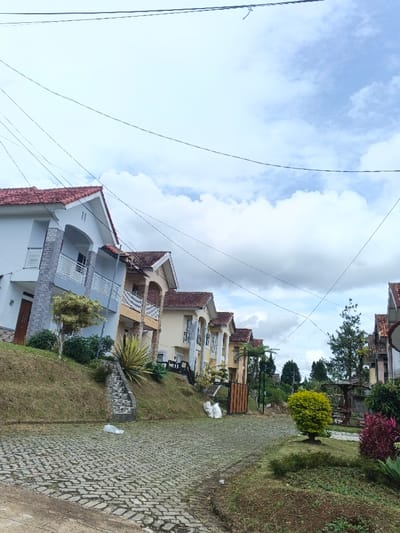 A row of hillside houses with red roofs and light-colored facades lines a cobblestone street. Green grass and various plants adorn the landscaped yards in front of the homes. Overhead, a network of power lines crisscrosses against a cloudy sky. The scene evokes a sense of quiet suburban living in a hilly environment.