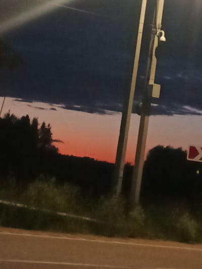The image captures a dusky sky with a gradient of dark blue to pale orange over a silhouetted landscape of trees and vegetation. A utility pole, with what appears to be a security camera mounted on it, stands prominently in the foreground, bisecting the scene vertically. A traffic sign, possibly indicating a turn, is visible on the right side. The lower portion of the image shows a road with white lane markings. The scene suggests an outdoor setting, possibly on the outskirts of Воскресенское, Russia, during twilight.