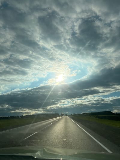 The image depicts a view from inside a vehicle looking forward onto a two-lane asphalt road. The road stretches towards the horizon, flanked by green fields on both sides. The sky is filled with dramatic, heavy clouds, with the sun breaking through in the upper center, casting bright rays of light. A few vehicles are visible in the distance on the road. The overall scene suggests a drive through a rural or semi-rural landscape, possibly during late afternoon or early evening, given the lighting. There is no specific indication of the city of Сухая Балка, Russia, in this image.