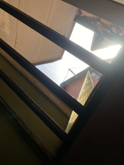 The image is taken from beneath a skylight-like structure composed of rectangular frames with some frames appearing to contain translucent material or screens. The structure appears old and possibly weathered. Outside, there's a view of buildings with a reddish-brown roof visible through one of the framed sections. The sunlight is bright, possibly indicating a clear day. No people are visible in the image. The image doesn't provide enough detail to confirm the exact location in Oke Ogun, Nigeria.