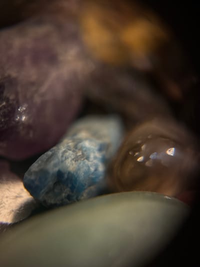 The image is a close-up, macro photograph of small, irregularly shaped stones, possibly gemstones or decorative gravel. The stones vary in color, including shades of purple, blue, and brown, with some appearing translucent and reflecting light. The background is dark and out of focus, creating a shallow depth of field that emphasizes the textures and details of the stones. There are no people or discernible actions depicted. The image does not contain any elements that would allow for the identification of a specific location within Biržai, Lithuania.