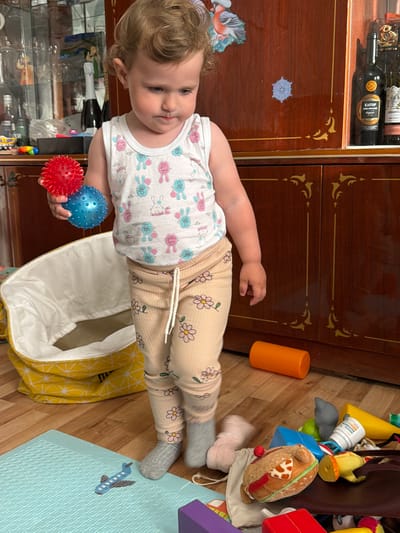 A toddler stands on a wooden floor in front of a wooden cabinet. The toddler is wearing a white tank top with cartoon rabbits and beige pants with a floral pattern, and grey socks. They are holding a red and blue textured ball in their left hand. To the left of the toddler is a large, yellow and white patterned laundry basket. Scattered on the floor are various toys, including a blue airplane toy on a light blue mat, an orange cylinder, a stuffed hamburger, and other colorful objects. The wooden cabinet behind the toddler has decorative gold trim and appears to be part of a room. The scene is indoors, in what looks like a living area or playroom. The location is Komsomolskiy, Russia.