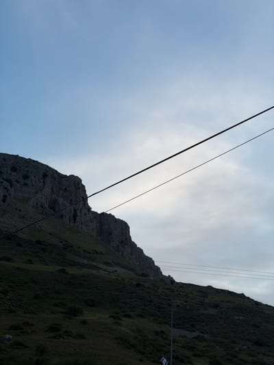 The image displays a steep, rugged mountain slope against a cloudy sky, likely captured from an ascending cable car. The sky, occupying the upper-left portion, exhibits pale blue tones blending into brighter white cloud cover towards the right, suggesting an overcast or partly cloudy day with the sun potentially obscured.
The lower-right of the image is dominated by the mountain, which features distinct rocky outcrops and is heavily covered in dark green vegetation, including grass and shrubs. Multiple dark cables crisscross the scene; two thicker, prominent cables run diagonally across the sky and mountain, indicative of a cable car system. Several thinner, parallel cables are also visible along the mountain's vegetated slope, possibly part of the lift infrastructure or power lines.
On the right side of the mountain, a vertical pole-like structure with a horizontal arm is visible. From this arm hangs a blue and white flag, clearly identifiable as the flag of Israel due to its two blue stripes and the central Star of David design, a strong location cue for Migdal, Israel.
No people are visible. The overall scene depicts a natural, mountainous landscape during daytime, viewed from a low angle, highlighting both the natural terrain and human-made transport infrastructure.