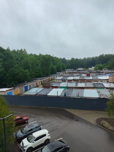 The image depicts a large, multi-row complex of garages situated in an urban environment, likely in Kaluga, Russia. The garages are constructed from various materials, including brick and corrugated metal, with predominantly grey, blue, and red doors, and a mix of metal and tar roofs. Many of the roofs show signs of wear and tear, with some appearing patched or degraded. Behind the garages, a dense line of deciduous trees forms a natural backdrop. In the foreground, a paved parking area is visible, wet from rain, with several cars parked: a red older model sedan with its roof open, a dark grey SUV, a white SUV, and a black sedan. The sky is overcast and grey, suggesting inclement weather. A dark grey metal fence separates the parking area from the garage complex. The overall impression is one of a utilitarian and somewhat aged urban development.