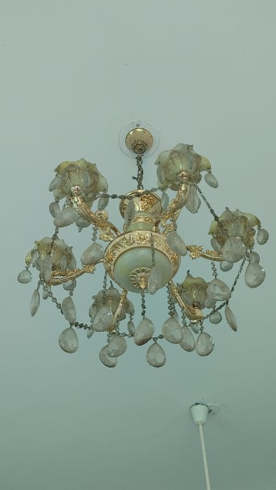 The image displays an ornate, multi-arm chandelier suspended from a light-colored ceiling. The chandelier features a central, light-toned body with intricate gold-colored decorative banding and finials. Six curved, gold-toned arms extend outwards, each supporting a light-colored, flower-shaped glass or alabaster cup. Numerous clear, faceted crystal pendants, predominantly teardrop-shaped, are suspended from the chandelier's arms and central structure by small chains.
The ceiling is a uniform light hue, possibly white or off-white, showing minor dust or discoloration around the chandelier's circular mounting plate. In the lower right portion of the frame, a separate white cylindrical ceiling fixture, possibly a light cord or fan mount, is visible, also with some discoloration at its attachment point. The scene is an interior environment, consistent with a residential or commercial building. No individuals, specific actions, or interactions are depicted. This scene is located within the city of Savar, Bangladesh.