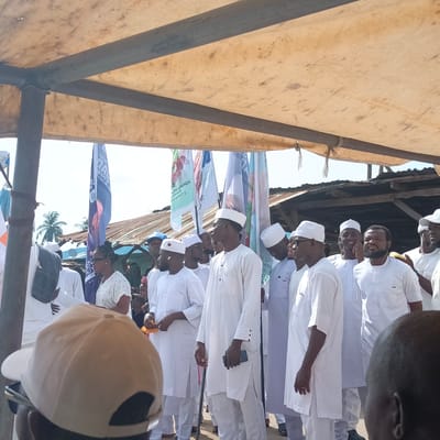 The image shows a group of people gathered under a canopy in Gudi, Nigeria. Most of the men are wearing white traditional attire, including long robes and caps. A few women are present, dressed in western clothes. Several flags are visible in the background, indicating a political rally or gathering. The environment appears to be a semi-urban area, with buildings and trees visible in the background. Some people are holding cellphones and other small objects. The overall scene suggests an event or meeting taking place in an open-air setting.