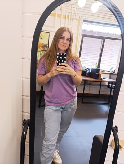 A woman is taking a selfie in a full-length mirror. She has long, blonde hair and is wearing a purple t-shirt and light blue jeans. She is holding a smartphone with a daisy-patterned case in her right hand. She has two rings on her right hand and a bracelet on her left wrist. She is smiling faintly. The mirror is framed in black and has an arched top.

The background reflected in the mirror shows an office-like setting. There is a desk with a computer monitor, keyboard, and other office supplies. Behind the desk, there are windows with horizontal blinds. There is also a painting on the wall to the left of the woman. The floor appears to be tiled. The lighting suggests it is daytime. The location is Chernogolovka, Russia.
