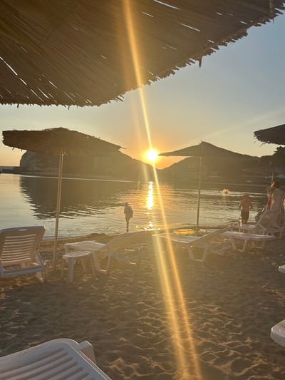 This is a photograph of a beach in Kavarna, Bulgaria, during sunset. The sun is low on the horizon, casting a warm, golden light across the scene. The beach is made of sand, and there are several white lounge chairs and a small table set up, suggesting a tourist resort. In the foreground, the straw roofs of two beach umbrellas are visible, with one umbrella prominently featured. The sea is calm, with gentle waves lapping at the shore. In the distance, a rocky cliff face with buildings is visible, and the sun's reflection shimmers on the water. There are a few people in the water, including what appears to be two children. One child is standing near the shore, and another is further out in the water. A woman is standing near some lounge chairs, and another person is partially visible behind her. The overall atmosphere is peaceful and serene, capturing the beauty of a sunset at the seaside.