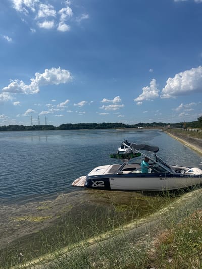 The image shows a wakeboard boat docked on the shore of a body of water, likely a canal given the straight banks and visible power lines in the distance. The boat is white with a dark blue stripe and the "MasterCraft" logo is visible on the side. A green and black wakeboard is leaning against the boat's wake tower. A person wearing a teal shirt is seated in the boat. The water is calm and reflects the blue sky, which is dotted with fluffy white clouds. The shore is lined with dry grass and some greenery. The overall scene suggests a sunny day, possibly late morning or early afternoon, ideal for water sports. The location is identified as Altenheim, Germany. Visible text includes "MasterCraft", "X2", "USA", and a boat registration number "SG DH 181". The presence of power lines and the engineered canal suggest an industrial or recreational area.
