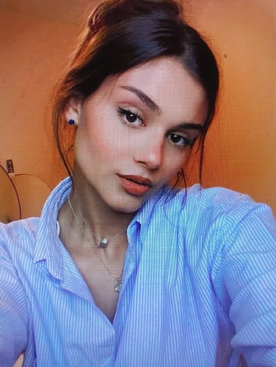 A close-up portrait features a young woman with dark hair pulled back in a bun. She is wearing a light blue striped button-down shirt and a necklace with two pendants. She has on subtle makeup, including eyeliner and lipstick. A small blue earring is visible in her left ear. The background is a solid, warm orange color, suggesting an indoor setting with warm lighting. There is no identifiable urban environment or specific location in Sluck, Belarus, visible in the image.
