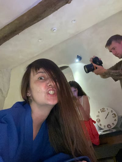 The image shows an indoor scene with three people. In the foreground, a woman in a blue robe is looking at the camera with her mouth open, making a funny face. Her hair partially covers her face. In the background, a man is holding a camera and appears to be taking a photo of a woman who is partially obscured by the foreground woman's hair. There is a round clock on a shelf behind the woman with the camera. The ceiling has exposed wooden beams and what appear to be smoke detectors. The scene is dimly lit. The location is not specified in the image or context provided.