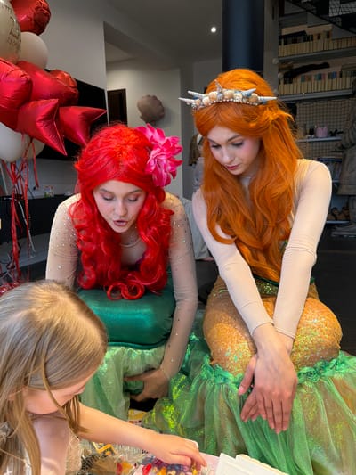 In a bright indoor setting, two individuals costumed as mermaids are positioned vertically, attending to a child visible in the lower right corner. The upper mermaid has long, red-orange hair adorned with a seashell crown, and wears a shiny gold and green mermaid tail. The lower mermaid has bright red, curly hair with a large pink flower, and a shiny green mermaid tail. Both are wearing light beige, long-sleeved tops with small embedded gems. To the left are several red heart-shaped and white round balloons. The location is Крючково-2 Снт, Russia.