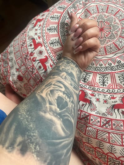 The image shows a close-up of a person's arm and hand resting on a pillow with a red and white patterned fabric. The arm is extensively tattooed with a dark, abstract design. The hand has long, almond-shaped nails painted with a French manicure. The pillow's pattern features circular motifs and stylized figures resembling horses or birds in red and black on a white background. The environment appears to be indoors, with soft lighting. The location of Коробицыно, Russia, is not discernible from the provided image.