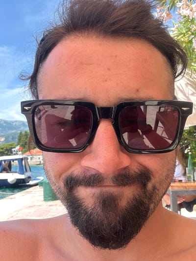 A close-up selfie-style portrait captures a man from the chest up, centered in the frame. He has dark brown, disheveled hair and is wearing black, square-framed sunglasses with dark lenses, which bear the "POLICE" brand logo on the right lens. His forehead shows tanned skin with minor blemishes and a slight scar-like mark on the left side (viewer's left). A short, dark brown beard covers his chin and jawline, and his nose appears tanned. His bare shoulders and neck are visible, also tanned, with visible goosebumps.
The sunglasses reflect the surrounding environment. The left lens reflects a blue body of water, a white boat with a blue canopy docked at a stone pier, distant green trees, and red-roofed buildings against a backdrop of mountains under a clear blue sky. The right lens reflects an outdoor dining setting, including a wooden table with metal legs, a glass bottle, and the legs of another person seated nearby, under what appears to be a white umbrella or canopy.
In the background, behind the man's left side (viewer's left), a tranquil blue bay is visible with a white boat docked at a stone pier. Lush green vegetation and traditional red-roofed buildings line the distant shore, backed by mountains. On the man's right side (viewer's right), flowering pink and white oleander plants and a white structure, likely part of an outdoor establishment, are visible under a bright, sunny sky. The scene is consistent with a coastal setting in Luštica, Montenegro.