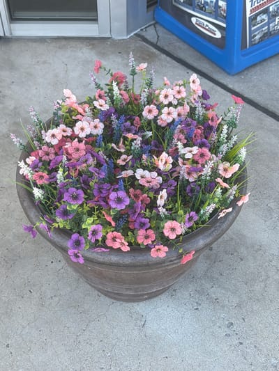 A large, round, dark brown planter filled with artificial flowers in various shades of pink, purple, and white sits on a gray concrete surface. The flowers are densely packed and include delicate, star-shaped blooms with darker centers, as well as spike-like clusters of lavender and tiny white flowers. Green foliage is interspersed throughout. In the background, a light-colored building with a horizontal window is visible, along with a blue stand displaying promotional material, possibly for a trucking publication, with the words "Truck Paper" clearly legible. The concrete surface shows a cracked texture and a dark line running across it. The scene appears to be outdoors, likely in front of a business. The location is the city of Lebanon, United States of America.