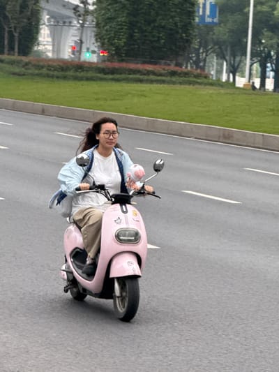 A woman rides a pink scooter on a road in Shuangfu, China. The woman has dark hair, wears glasses, a white t-shirt, tan pants, and a light blue jacket. She is holding a small pink object in her left hand. The road has white lane markers and is bordered by a grassy median. Traffic signals are visible in the background.
