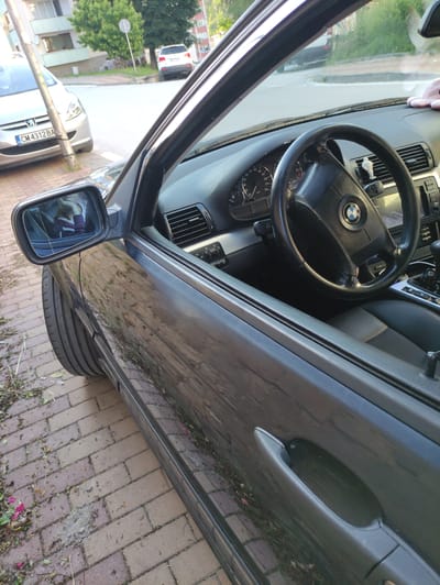The image shows the interior and exterior of a dark-colored BMW parked on a brick sidewalk. The driver's side door is open, revealing the steering wheel and dashboard. A person is visible in the side mirror, wearing a purple and white top. Outside the car, a silver Peugeot with a license plate reading "CM 4312 BA" is parked behind it. In the background, a white SUV is visible on a street, with buildings and trees lining the area. The overall scene appears to be a street in a residential area of Madan, Bulgaria.