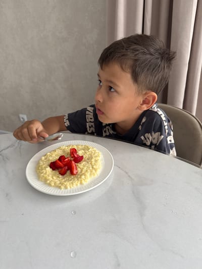 A young boy sits at a marble table in an interior space. He is looking to his right with a bowl of oatmeal topped with sliced strawberries in front of him. He is holding a spoon in his right hand, poised as if to take a bite. His dark hair is short and tousled. He is wearing a dark t-shirt with a graphic print. The background shows a textured wall and draped curtains. The image does not provide enough information to determine its location in Nizhnyarkeyevo, Russia.