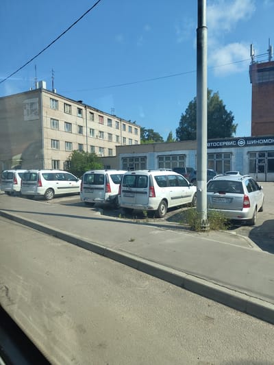 The image depicts a street scene in the city of Zhukovsky, Russia, on a clear day. A light gray, multi-story apartment building stands on the left side of the frame, its brick facade and numerous windows visible. To the right of the apartment building, a low-rise commercial building with a blue and gray facade is visible, featuring several large windows and signage that appears to read "АВТОСЕРВИС" and "ШИНОМОНТАЖ", indicating an auto service and tire fitting shop. A tall, gray utility pole stands prominently in the foreground. Parked along the sidewalk in front of the commercial building are several white Lada Granta station wagons. One silver Chevrolet Aveo is also parked in the vicinity. The street and sidewalk are paved, with some patches of grass growing through cracks. The sky above is a clear, vibrant blue.