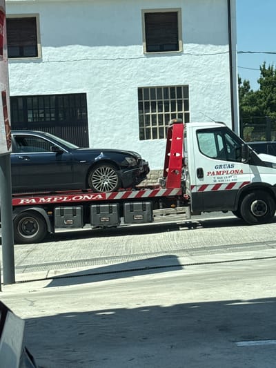 The image shows a dark blue sedan with significant front-end damage, including a crumpled bumper and hood, being transported on a tow truck. The tow truck has "GRUAS PAMPLONA" printed on its side, along with phone numbers. The truck is white with red and white striped safety markings. The tow truck is parked on a paved surface in front of a white stucco building with multiple windows. The building appears to be a warehouse or industrial facility. Trees are visible in the background. The scene suggests the vehicle is being towed for repairs in or around Pamplona, Spain. The lighting indicates it is daytime, likely sunny.