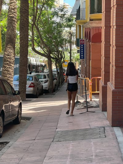 A woman with long dark hair, wearing a white top and dark shorts, walks away from the camera on a sidewalk. She is seen from the back, and her legs are visible. Behind her, a row of cars is parked along the street. Trees line the sidewalk, casting shadows. The buildings in the background are made of brick and have colorful facades. A blue sign with a Union Jack and the word "ACADEMY" is visible on the left. The scene appears to be an urban environment on a sunny day.