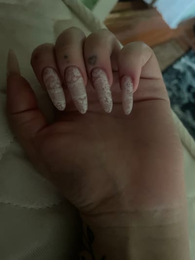 In the city of Linëvka, Russia, a close-up shot captures a person's hand with long, almond-shaped nails adorned with white lace patterns. The nails are a light pink base with intricate white lace designs, creating a delicate and feminine look. There are visible tattoos on the fingers, including a small dot on the middle finger and a heart shape on the ring finger. The hand is resting on a textured surface, possibly bedding or upholstery, which appears to be a light color. The background is softly blurred, showing hints of wooden furniture and a dark, textured fabric, suggesting an indoor setting. The lighting is dim, casting soft shadows and highlighting the details of the nails and skin.