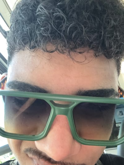 The image shows a close-up, low-angle, selfie-style photograph of a person's face, from the forehead down to the chin. The individual has dark, curly hair and is wearing large, green-framed sunglasses that obscure their eyes. A hint of a mustache is visible above the upper lip. The background is blurred and appears to be indoors, possibly a vehicle, with some indistinct shapes suggesting a window and interior elements. The lighting is bright, coming from above and to the side. The photograph does not provide any identifiable details of Kazichene, Bulgaria.