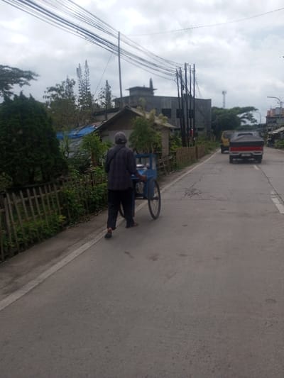 An outdoor scene in Sarwahi Mal., India, depicts a man walking away from the viewer on the left side of a paved asphalt road. The man wears a plaid shirt, dark pants, and a cap, pushing a blue two-wheeled cart. A solid white line marks the immediate left edge of the road. Adjacent to the road, a wooden picket fence borders an area of dense green foliage, including a prominent conical evergreen tree and smaller structures, one featuring a light-colored roof and another covered by a blue tarp. Overhead, multiple black utility lines span across the frame, supported by several utility poles on the right side of the road. In the mid-ground, a large, grey, uncompleted concrete building with visible window openings is present. Further down the road, two vehicles, a yellow truck followed by a red truck, are observed moving away from the viewer. A cellular tower is visible in the far distance on the right. The sky is overcast with light grey clouds.