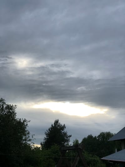 A low-angle, medium shot captures a view of a cloudy sky above a dense treeline and a partially visible building in the foreground. The sky is dominated by thick, grey clouds with a bright, irregular patch of sunlight breaking through near the center. The clouds appear layered and textured, with some lighter areas suggesting the sun's rays diffusing through them.

Below the sky, the upper branches of dark green deciduous trees fill the left and central portions of the frame. To the right, a tall, dark evergreen tree stands out, its needles finely detailed. Further to the right, the dark grey, sloped roof of a building is visible, its corrugated texture apparent. A wooden swing set structure with rough-hewn beams is positioned in the lower-middle section of the image, partially obscured by foliage. The overall lighting suggests either early morning or late evening conditions, with the bright patch in the sky creating a dramatic contrast against the darker elements. The scene is devoid of human presence or any apparent activity. The location is identified as Zhukovo, Russia.