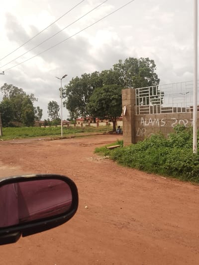 The image captures a scene in Bokkos, Nigeria, featuring a dirt road in the foreground. A vehicle's side mirror is visible on the left, reflecting a reddish surface. To the right, a brick wall with graffiti reading "ALAMS 2021 DP" stands, partially obscured by lush green vegetation. Beyond the wall, a white metal fence with an abstract design encloses an area. In the background, a solitary figure sits under a tree, and a street light stands near the fence. Further back, trees and buildings line the horizon, under a cloudy sky. The overall environment suggests a semi-urban or suburban setting.