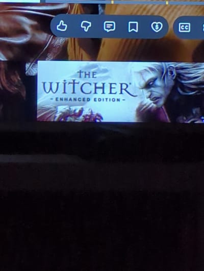 The image shows a screenshot of the video game "The Witcher: Enhanced Edition" displayed on a screen. The game's title is prominently visible in large, stylized font, with "THE" above "WITCHER" and "- ENHANCED EDITION -" below. A portion of the game's artwork, featuring a character with pale skin and long white hair, is also visible. Above the game title, a dark bar with several icons is displayed. These icons appear to be related to media playback or user interface elements, including a thumbs-up, thumbs-down, a speech bubble, a bookmark, a heart with a dollar sign, and a "CC" symbol. The background of the screenshot is blurred and appears to be a brown, textured surface. The image is taken from a slight angle, with a dark area at the bottom, possibly indicating the lower portion of the screen or the surrounding environment. There is no visible indication of the city of Erlangen, Germany, in this image.