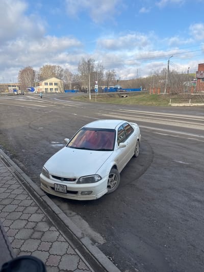 A white Honda Euro R coupe is parked on the side of a street in Mesyagutovo, Russia. The car is situated on a patch of asphalt adjacent to a sidewalk made of patterned grey and white pavers. The street surface shows signs of wear and discoloration. In the background, there is a view of trees without leaves and a building with multiple windows. Blue utility structures are visible along the roadside, and power lines stretch across the sky. The sky is partly cloudy. The license plate is partially obscured but appears to read "T 745 EK 102". There are no people immediately visible near the car. The environment suggests an urban setting in a state of disrepair.