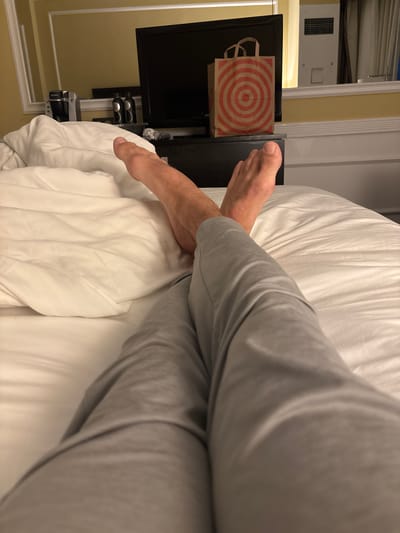 A first-person perspective shot shows a person's legs and feet resting on a white bed. The person is wearing light grey pants. The background features a television set with a red and white bullseye patterned bag on top of it, a coffee maker, and a mirrored wall reflecting a window and a heating unit. The scene appears to be in a hotel room in San Francisco, California, United States of America.