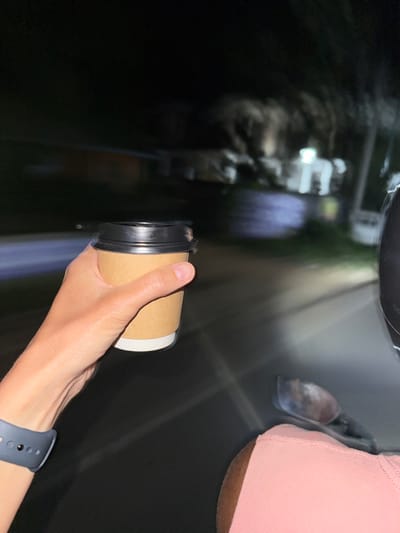 At night in Dikwella, Sri Lanka, a right human hand is depicted holding a standard disposable beverage cup. The cup is light brown with a white lower section and a black plastic lid. A dark blue wristband, consistent with a smartwatch or fitness tracker, is visible on the individual's left wrist in the lower-left corner. A portion of a light pink garment, likely worn by the same individual, is visible on the right side of the frame, below a dark-colored helmet. A blurred metallic object, possibly a vehicle rearview mirror or bodywork, is present below the helmet. The scene is observed from a moving vehicle, indicated by motion blur in the background. The environment is dark, with indistinct, blurred shapes of trees or foliage and distant artificial lights. A single prominent light source illuminates a section of the asphalt road ahead, casting a glare.