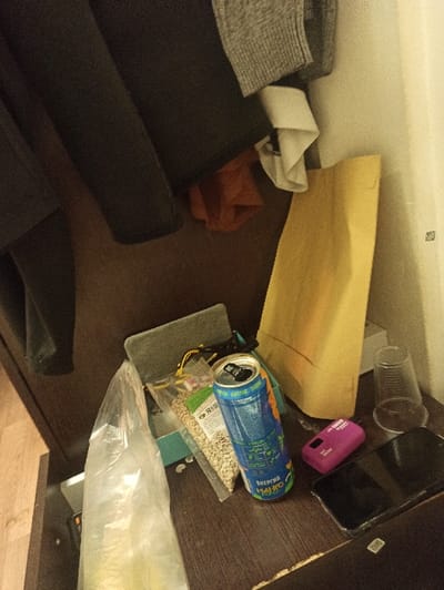 The scene depicts a cluttered interior, specifically a tabletop or shelf within a room at a Tecnimont camp in Russia. In the upper left, various articles of clothing are visible, including dark trousers, a grey knitted garment, a white collared shirt, and an orange fabric piece, hanging against a dark wooden surface. A light-colored wall segment is visible on the upper right.

The dark wooden tabletop in the foreground holds several items. From left to right: a translucent plastic bag, partially filled; behind it, a clear plastic bag containing numerous beige seeds, likely sunflower seeds, with a label displaying "SPS" in Cyrillic. Adjacent to these, an opened blue and green energy drink can, featuring "ЭНЕРГИЯ МАНГО" (Energy Mango) in Cyrillic script, stands upright. Further right, a yellow paper bag stands. Next to the bag, an empty, clear disposable plastic cup is present. To the right of the cup, a magenta-colored portable power bank lies beside a black smartphone, which is face-up with its screen dark, encased in a clear protective cover. No individuals are present in the image.