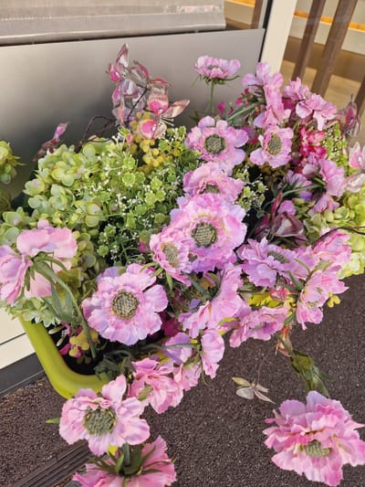 A bright green planter holds a vibrant arrangement of artificial flowers, predominantly in shades of pink and light green, accented by small yellow blooms and delicate artificial butterflies. The planter is situated outdoors on a dark, textured surface that appears to be pavement or gravel. In the background, a building with horizontal wooden slats and a reflective, metallic surface can be seen. The scene suggests an exterior decoration likely placed at the entrance of a commercial establishment or private residence. The location of Antony, France, is not visually indicated by any specific landmarks or context within the image.
