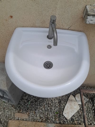 A white ceramic sink with a silver faucet is mounted on a textured, light-colored wall. The sink is empty and appears slightly damp, with water droplets visible inside. A drain with a metal grate is located at the bottom of the sink basin. The wall has some minor cracks and discoloration. To the left of the sink, a grey concrete block is visible, and to the right, the ground is covered with small grey pebbles. A piece of broken tile rests on the pebbles to the right of the sink. The scene is set outdoors, in what appears to be a utilitarian or outdoor washing area. The overall impression is one of simplicity and practicality.