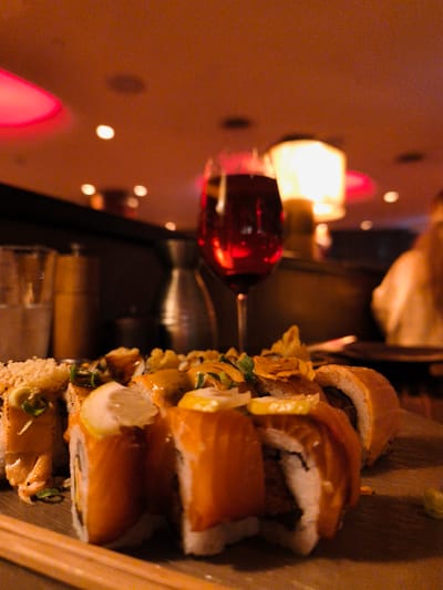A close-up, low-angle shot focuses on a platter of sushi rolls and a glass of red wine in a dimly lit restaurant. The sushi rolls are prominently displayed in the foreground, with some topped with what appears to be salmon and a creamy sauce, garnished with thinly sliced green onions and lemon wedges. Chopsticks rest horizontally in front of the sushi. Behind the food, a glass of red wine is visible, with its deep red color reflecting the ambient light. In the background, soft, warm lighting emanates from lamps, suggesting an evening setting. Out-of-focus shapes of restaurant patrons and decor can be seen, creating a sense of a lively, yet intimate, atmosphere. The overall mood is cozy and inviting, suggesting a pleasant dining experience. There is no visible text in the image.