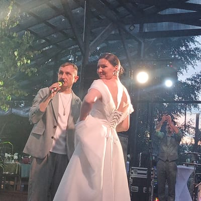 The image depicts an outdoor event with a focus on two central figures and a third person in the background. On the left, a man in a light grey suit and a white t-shirt is holding a microphone to his lips, suggesting he might be speaking or singing. He wears a necklace with a pendant. To his right, a woman in a white wedding dress, featuring an intricate corset-style back closure and flowing fabric, is turned away from the camera, looking over her shoulder with a smile. Her hair is styled up, and she is wearing large earrings. In the background, a man is holding up a smartphone, seemingly taking a picture or recording a video of the scene. The setting appears to be an outdoor stage or venue, possibly at dusk, as indicated by the lighting and the sky visible through the overhead structure. The atmosphere seems festive or celebratory. The background shows trees and foliage, hinting at a natural or garden-like location. There is also some audio equipment visible, such as speaker cases. No discernible text is present in the image.
