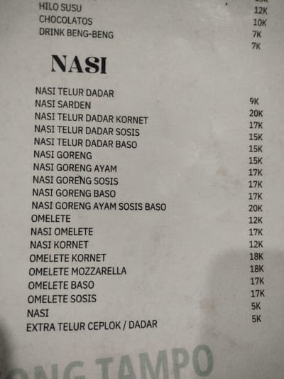 This media file is a photograph of a printed menu, likely from a casual eatery in Dramaga, Indonesia. The menu is printed on white paper with black text and appears slightly creased and aged. It lists various food and drink items with their corresponding prices.
The top visible section includes drinks:
- HILO SUSU (12K)
- CHOCOLATOS (10K)
- DRINK BENG-BENG (7K, followed by another 7K, suggesting two distinct "DRINK BENG-BENG" options or a repeat).
The main section, prominently titled "NASI," lists rice-based dishes:
- NASI TELUR DADAR (9K)
- NASI SARDEN (no price visibly associated)
- NASI TELUR DADAR KORNET (20K)
- NASI TELUR DADAR SOSIS (17K)
- NASI TELUR DADAR BASO (15K)
- NASI GORENG (15K)
- NASI GORENG AYAM (15K)
- NASI GORENG SOSIS (17K)
- NASI GORENG BASO (17K)
- NASI GORENG AYAM SOSIS BASO (20K)
Following the "NASI" section, there are omelette and other rice-related items:
- OMELETE (12K)
- NASI OMELETE (17K)
- NASI KORNET (12K)
- OMELETE KORNET (18K)
- OMELETE MOZZARELLA (18K)
- OMELETE BASO (17K)
- OMELETE SOSIS (17K)
- NASI (5K, likely plain rice)
- EXTRA TELUR CEPLOK / DADAR (5K)
The prices are indicated with "K," which typically stands for thousands (Rupiah) in Indonesia, aligning with the location context. Part of a larger text, "ONG TAMPO," is visible at the very bottom of the menu. No activity or specific time of day is discernible from the image.