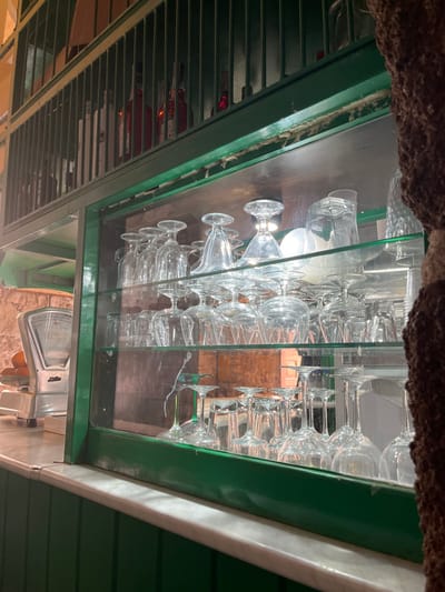 The image shows the interior of a bar or restaurant in Agüimes, Spain.  The location is near the coordinates latitude: 27.907, longitude: -15.447. The scene is captured from a low angle, looking up at a green-painted wall that has two shelves. The top shelf is a wire cage, and the bottom shelf is a glass-fronted cabinet.  The wire cage contains several bottles of liquor. The glass-fronted cabinet holds numerous empty glass cups and glasses. The lower shelf has a white marble countertop.  In the foreground, a silver scale sits on the countertop.  There are no people in the image.