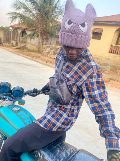 A man wearing a gray knit hat with cartoon eyes, a blue and red plaid shirt, black pants, and a dark-colored leather bag rides a light blue motorcycle. The background includes buildings and a palm tree. The scene appears to be outdoors during the day. The image is rotated.