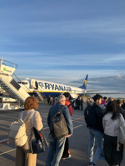 The image depicts a queue of passengers boarding a Ryanair airplane at an airport in Schönefeld, Germany. A mobile boarding stairs, labeled "WISAG," is positioned next to the aircraft. The plane is white with blue and yellow accents, and the Ryanair logo is visible on its tail and fuselage. Passengers, carrying luggage and backpacks, are seen walking up the stairs towards the plane's entrance. The scene takes place outdoors on an airport tarmac, with a clear blue sky and some clouds overhead. The lighting suggests it is either early morning or late afternoon.