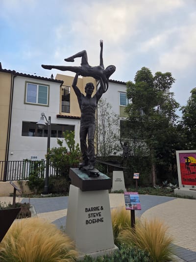 The image shows a bronze sculpture of a male and female figure, seemingly dancing or performing a lift, positioned in front of a modern building. The male figure is on the bottom, holding the female figure above him with outstretched arms. Both figures appear athletic and are depicted in motion. The sculpture is situated on a concrete pedestal that is inscribed with the names "BARRIE & STEVE BOEHNE".

In the background, there is a three-story building with a light-colored facade, featuring windows and balconies. To the right of the main sculpture, there is another, smaller bronze sculpture of a single figure also in motion, and further to the right, a colorful mural or poster is visible. The area around the sculptures is paved with patterned tiles and includes landscaping with ornamental grasses and shrubs. A lamppost is also present to the left of the main sculpture.

This scene is located at the Ocean Institute in Dana Point, California, United States of America. The Ocean Institute is a well-known marine science education center. The sculptures are part of an outdoor exhibit that celebrates individuals who have made significant contributions to surfing and ocean sports. The sculpture specifically depicts Barrie and Steve Boehne, renowned surfers and watermen. The overall environment suggests an outdoor public space, likely a plaza or walkway within the Ocean Institute grounds. The sky is overcast with some patches of brightness, indicating it is daytime.
