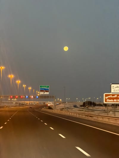 The image shows a roadway in Umm Salal Ali, Qatar at dusk or dawn. The moon is visible in the sky. Streetlights illuminate the side of the road. Traffic signs are visible.