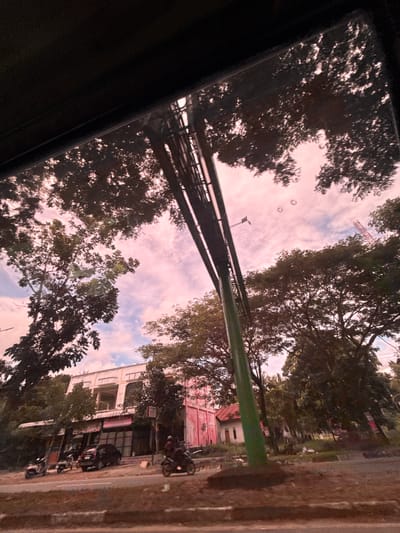 The image depicts a view from inside a vehicle, looking out at a street scene in Kampungblang, Indonesia. The sky is a soft pink, with scattered clouds. Trees with dense foliage frame the sky and a building complex. The building appears to be a commercial establishment with multiple storefronts and a pink facade on one section. Several motorcycles and a car are parked in front of the building, and a person is riding a motorcycle on the road. A tall green pole, part of some overhead infrastructure, is prominently featured in the foreground. The overall impression is of a busy, everyday street in a tropical environment, captured during what appears to be either sunrise or sunset due to the sky's color.