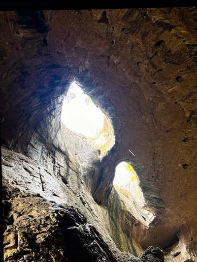 The image captures the awe-inspiring interior of a large natural cave, viewed from a low angle looking directly up towards its ceiling. Two distinct, irregularly shaped openings pierce the cave's roof, allowing brilliant natural light to flood into the cavern. These openings are famously known for their striking resemblance to a pair of human eyes, giving the cave its popular moniker, "The Eyes of God."
The rock walls surrounding these "eyes" are rugged, textured, and deeply grooved, showcasing layers and natural erosion patterns. The dominant colors are shades of earthy brown, grey, and dark ochre, with some areas appearing darker due to shadows and possibly moisture. The strong contrast between the brilliant white light streaming through the openings and the shadowy rock surfaces highlights the cavern's immense scale and dramatic contours. Hints of green vegetation are visible through the "eye" openings, suggesting an outdoor setting during daylight hours. No people, objects, or visible text are present. The scene is a powerful testament to natural geological processes, evoking a sense of wonder and grandeur. This formation is characteristic of Prohodna Cave in Bulgaria, aligning with the provided location context of Rosen, Bulgaria.