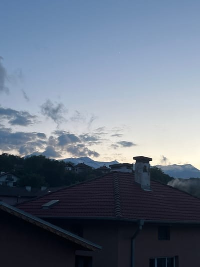 The image shows a view in Kornitsa, Bulgaria, featuring a sky with scattered clouds transitioning from blue to a lighter, almost white hue near the horizon. Mountains are visible in the distance, partially obscured by clouds and possibly fog. The foreground includes the roof of a building with red tiles and a chimney. Adjacent to the roof is part of a building with windows. The overall lighting suggests dawn or dusk.