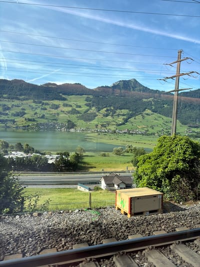 The image captures a scenic view from a train window, overlooking a tranquil lake, rolling green hills dotted with houses, and a majestic mountain range in the distance under a clear blue sky with wispy clouds. In the foreground, a section of railroad tracks and ballast is visible. To the right of the tracks, a wooden crate with green markings sits on the ballast. Beyond the tracks, a grassy field stretches towards a highway, with a few houses and dense green trees bordering the road. The lake is a central feature, reflecting the sky and surrounding landscape, with a village nestled along its shore. The distant mountains are covered in a mix of dark green forests and patches of lighter vegetation, suggesting a varied terrain. The overall impression is one of natural beauty and rural tranquility. The location is Steinen, Switzerland.